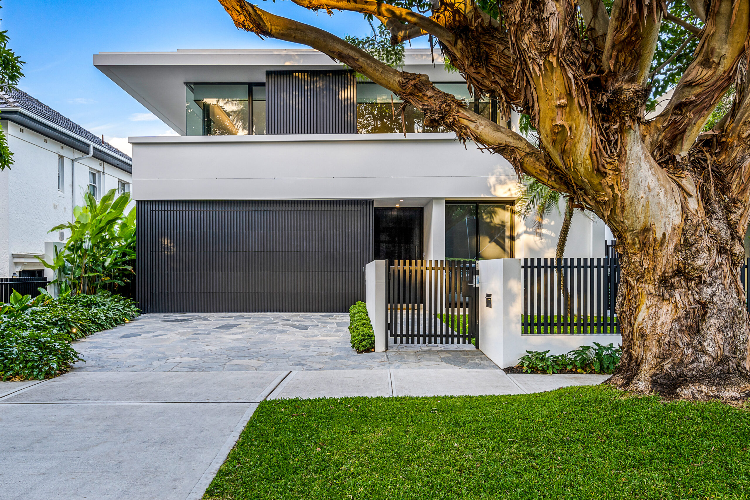 Modern home facade, featuring grey battens and floor to ceiling glass; softened with vivid green landscaping