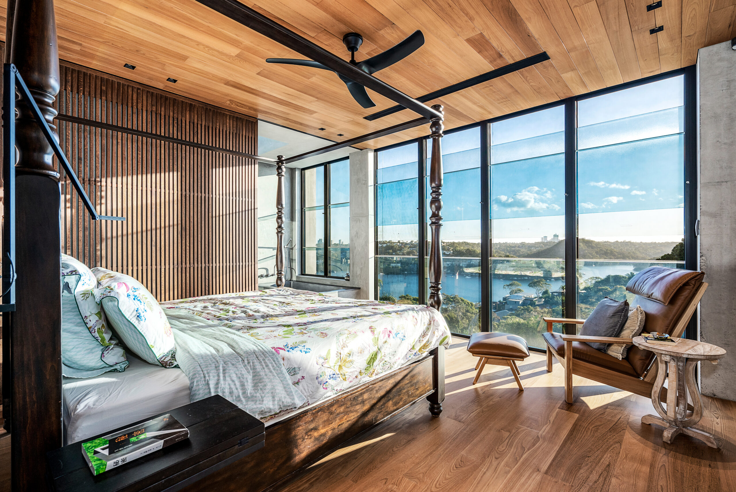 Four-poster bed with wooden paneled ceilings, carried through to batten walls and wooden flooring; looking onto a floor-to-ceiling water view
