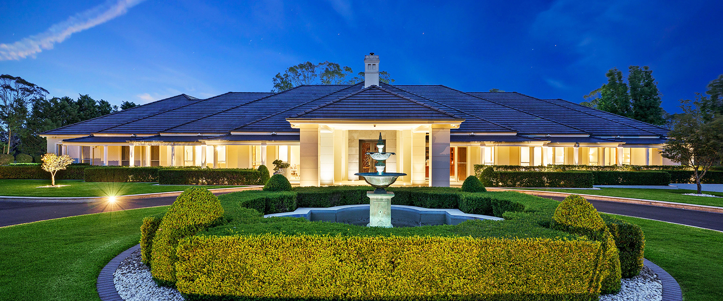 Palatial residence in Cobbitty, under a dark blue dusk sky; lit up with a warm glow, hedges and fountain centrepiece in the middle.
