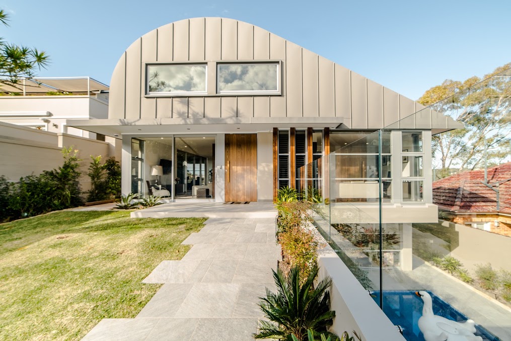 Multi-level contemporary house with a distinctive curved roofline. Grass to the left, and pool a level below.